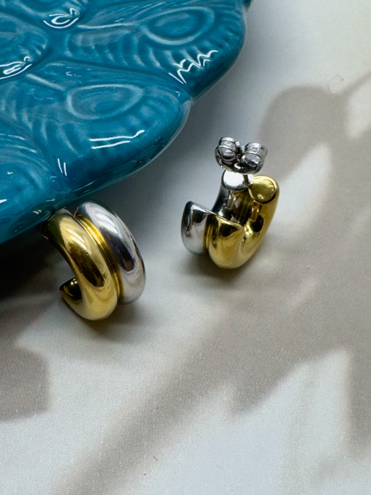 Gold and silver earrings on a textured white surface with a blue decorative object in the background.