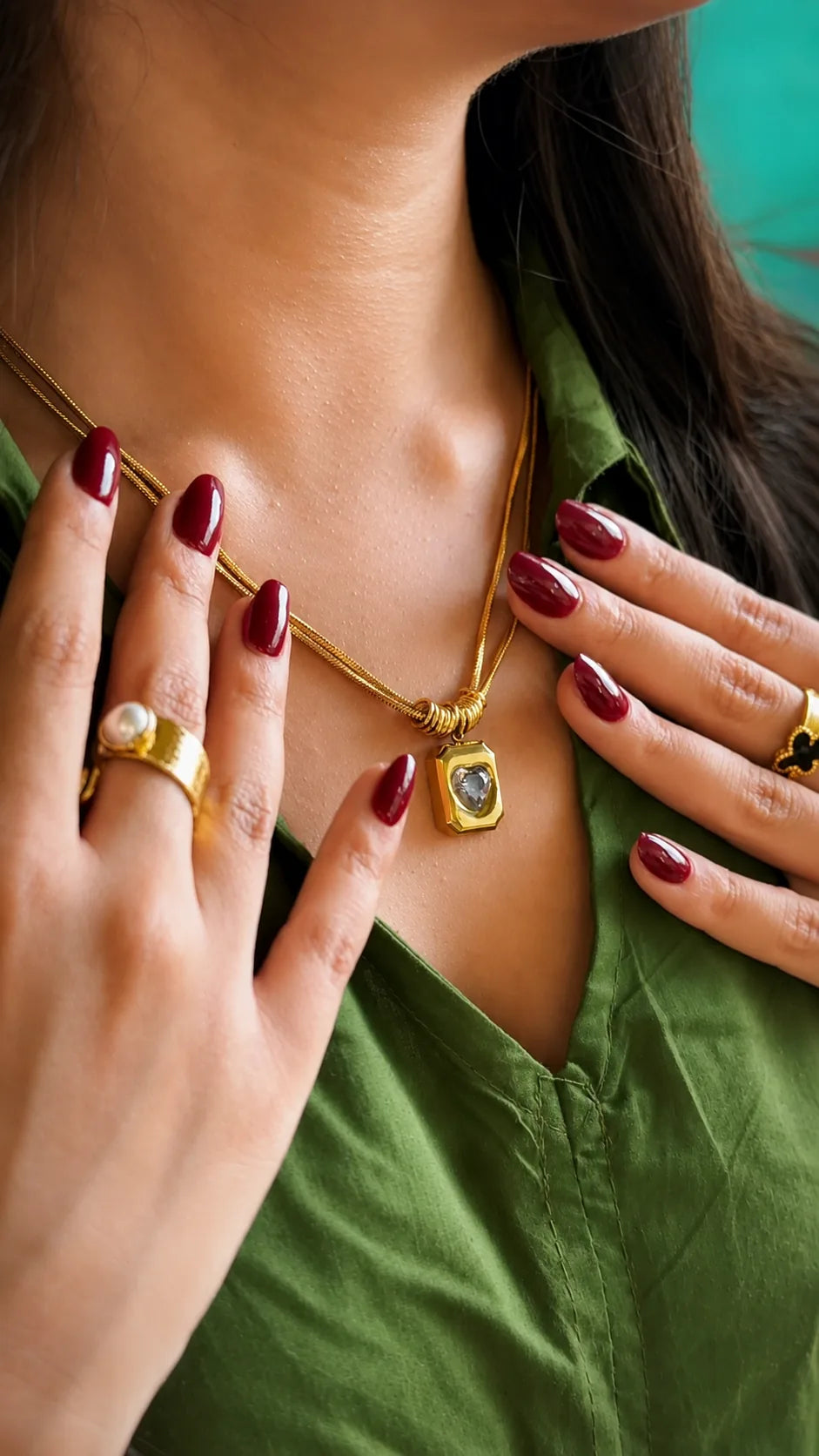 A close-up lifestyle shot of a woman wearing a gold chain necklace with a square pendant containing a clear teardrop-shaped gemstone. She is wearing an olive green shirt and has dark red manicured nails, with gold rings on her fingers.