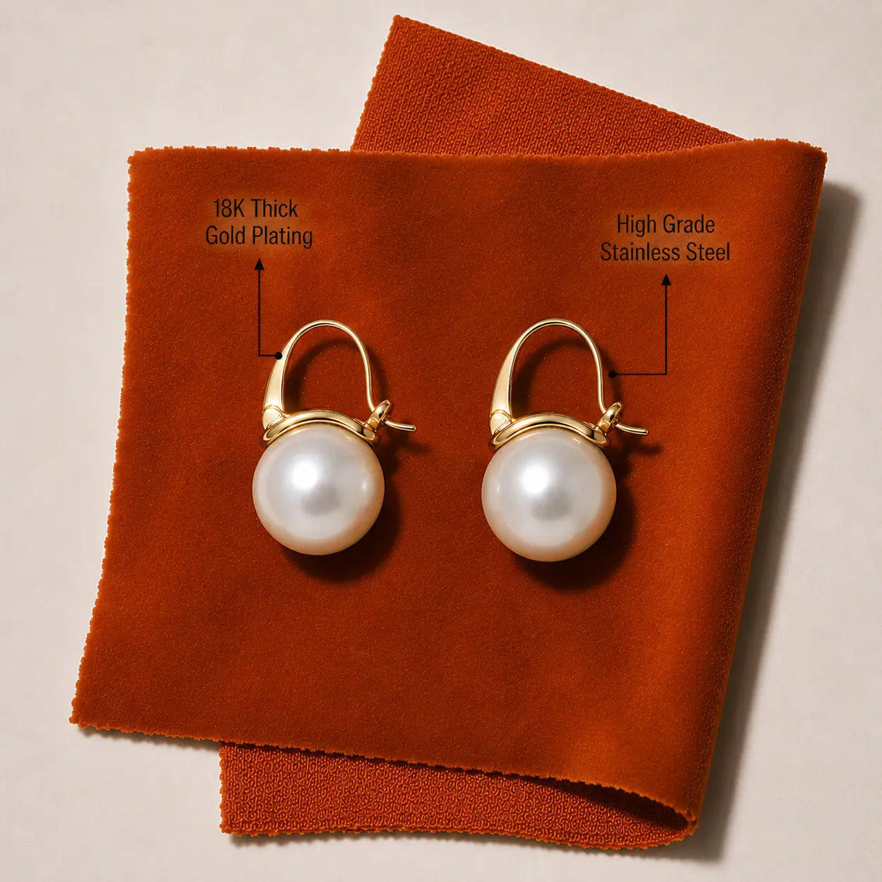 A pair of gold-plated and stainless steel hoop earrings with large white pearl drops, displayed on an orange-brown suede-like fabric background with text labels pointing to the materials.