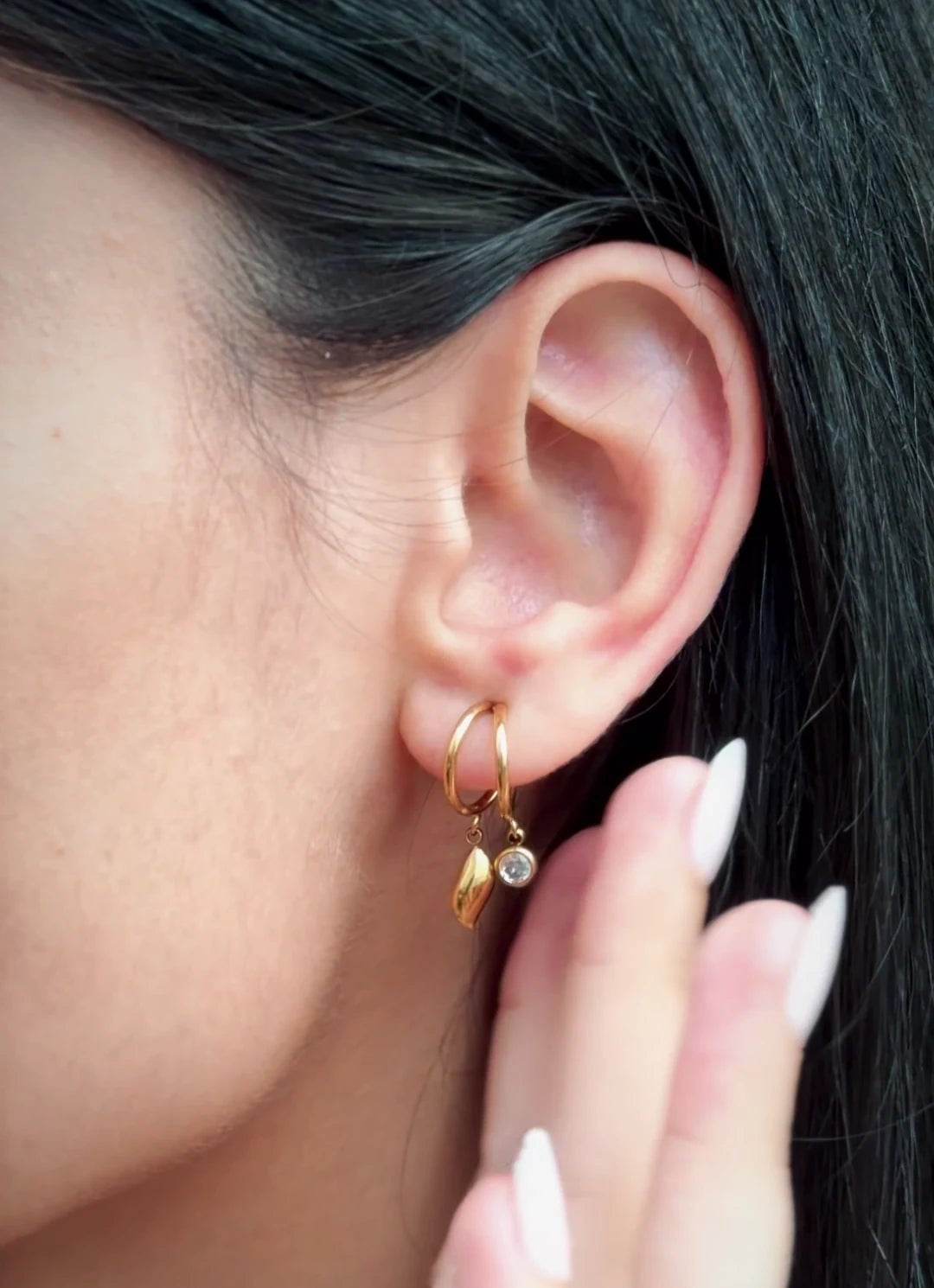 A close-up of a person's ear wearing a delicate gold hoop earring with small dangling gold charms, including a tiny clear stone accent. The person has dark hair and manicured fingernails.