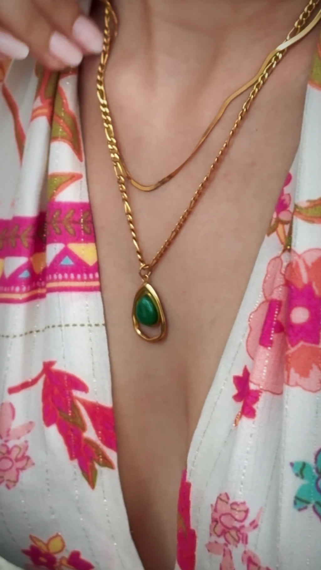 A close-up of a person wearing a layered gold necklace with a teardrop-shaped green stone pendant, paired with a white floral top.