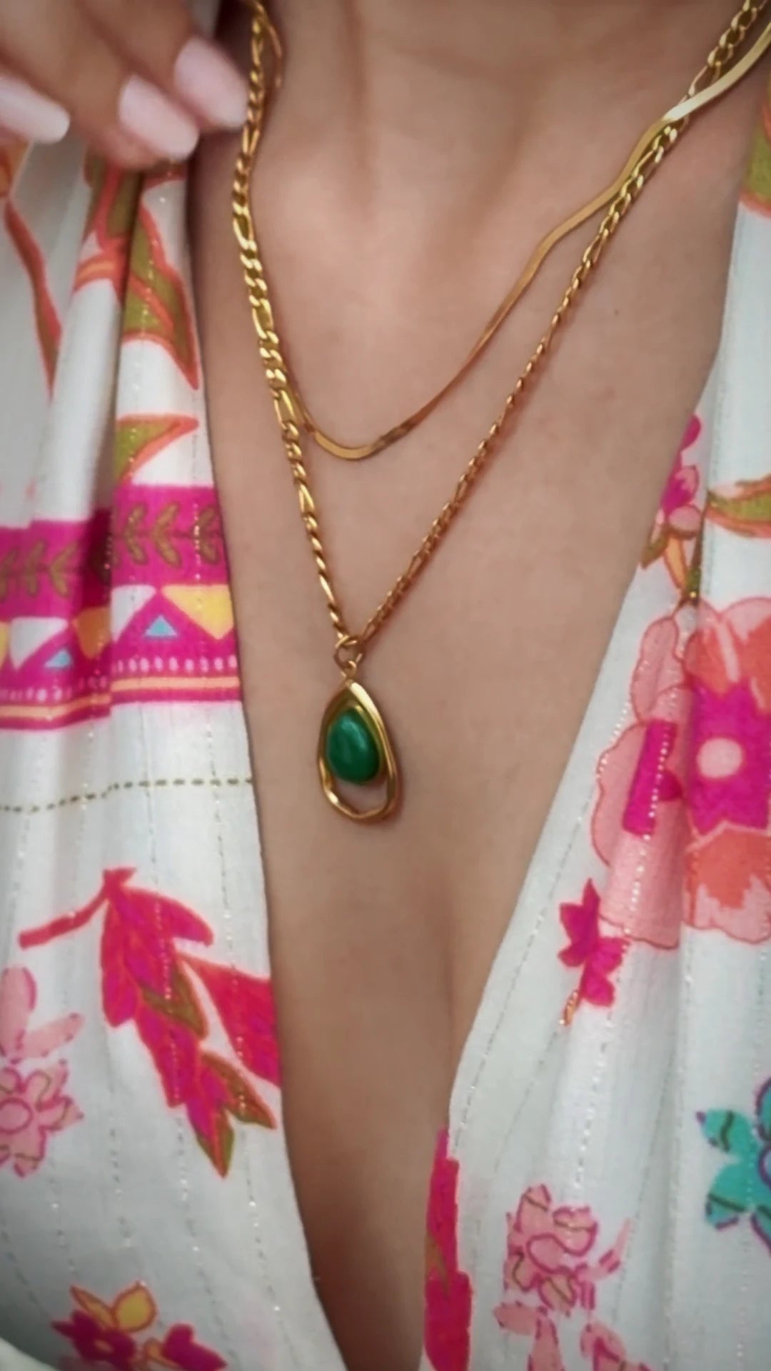 A close-up of a person wearing a layered gold necklace with a teardrop-shaped green stone pendant, paired with a white floral top.