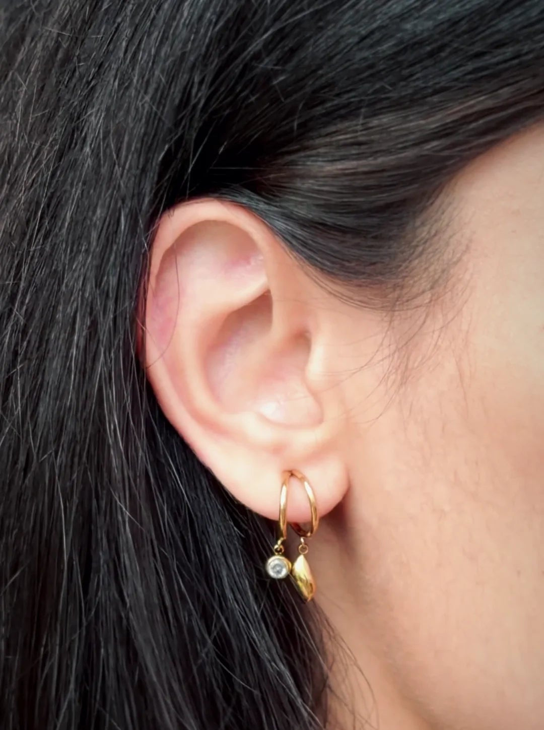 A close-up of a person's ear wearing a delicate gold huggie hoop earring with a small dangling gold leaf charm and a tiny clear stone accent.